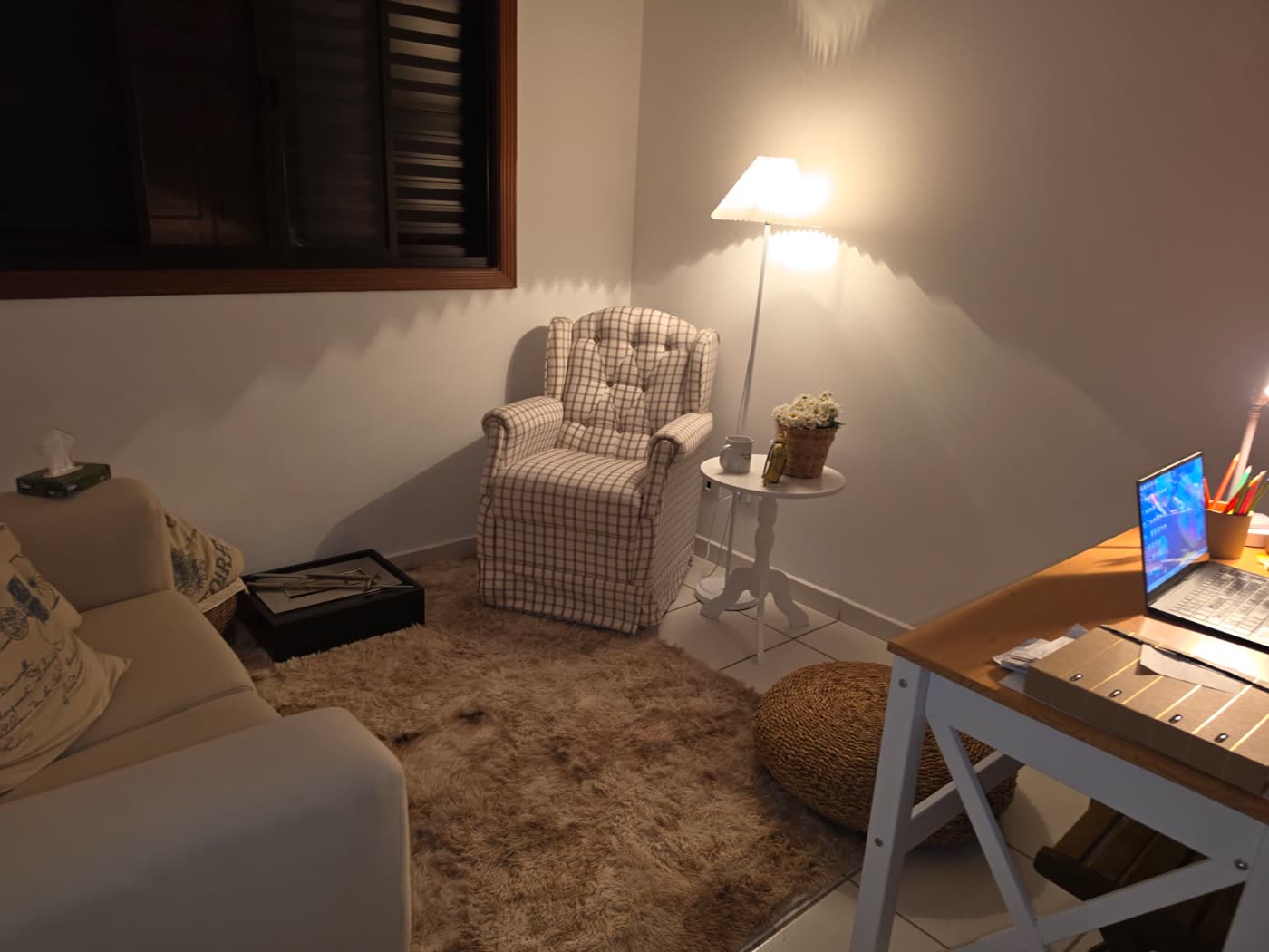 Sala de atendimento com poltrona xadrez, abajur aceso, sofá e tapete felpudo